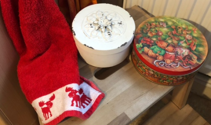 Ein hölzernener Hocker auf dem eine weiße und einen bunte, runde Keksdose stehen. Am Rand des Hockers ist ein rotes Handtuch drapiert, das eine weiße Borte hat auf der rote Rentiere abgebildet sind.